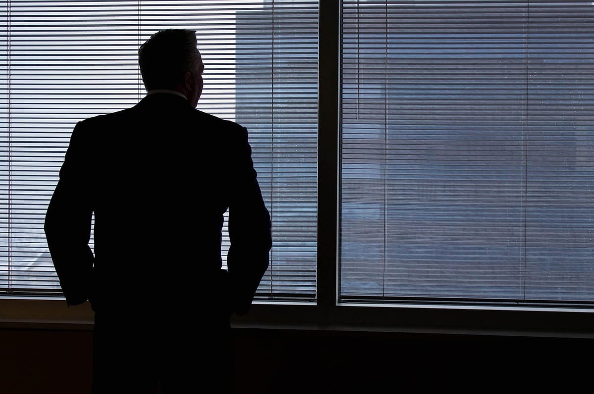 A man in a suit stands with his back to the camera, looking out through the blinds of a darkened office window.