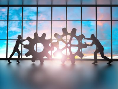 Two silhouetted business people push large gears together in front of a floor-to-ceiling window at sunrise.