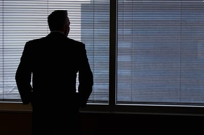 A man in a suit stands with his back to the camera, looking out through the blinds of a darkened office window.