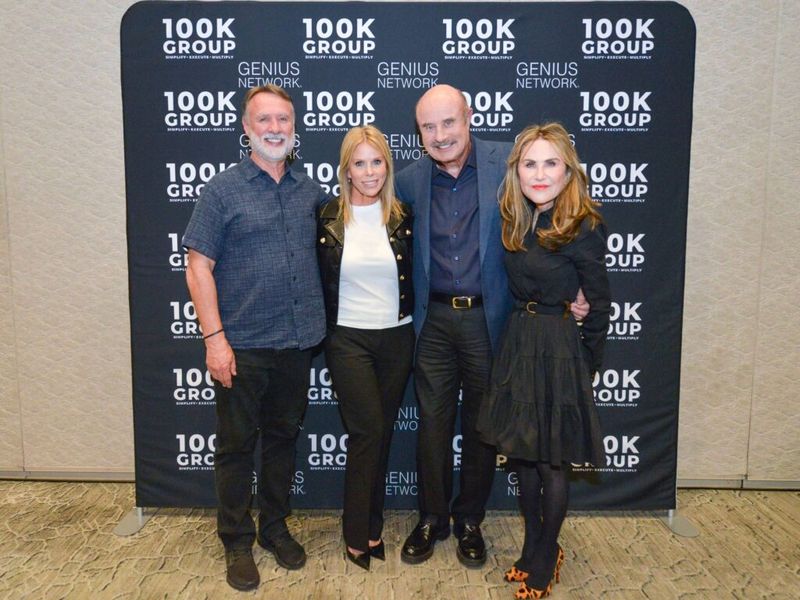 Randy Long posing with three others in front of a 100K Group and Genius Network step-and-repeat banner at a conference event
